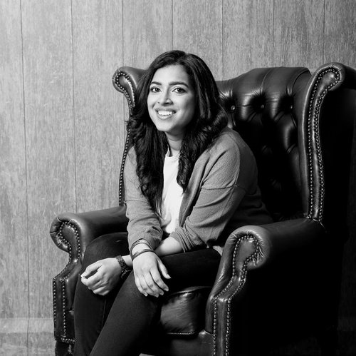 A black and white headshot of Archana Ramesh, who is sitting on a leather chair and smiling at the camera,