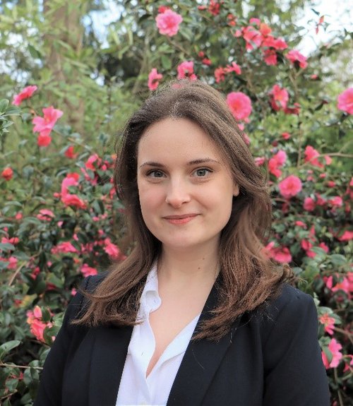 A headshot of Dr Eleanor Baker standing in front of a bush of pink flowers and smiling at the camera