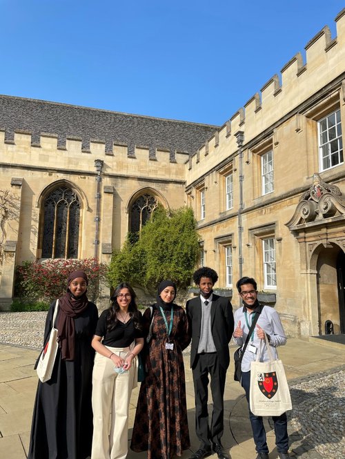 Five secondary school pupils stand in the St John&#x27;s front quad on a sunny day smiling. One of them holds a tote bag with the St John&#x27;s logo on it and does a thumbs up to the camera