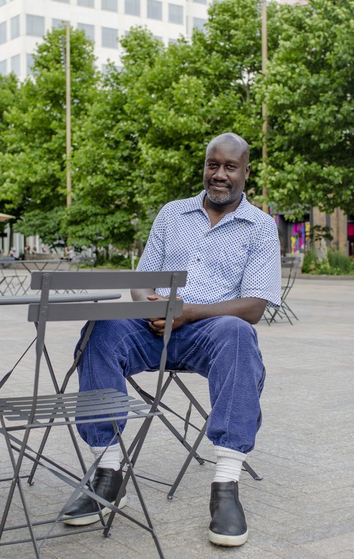 Musa Okwonga sits at an outdoor cafe table and looks into the camera with a slight smile. Trees and buildings can be seen behind him.